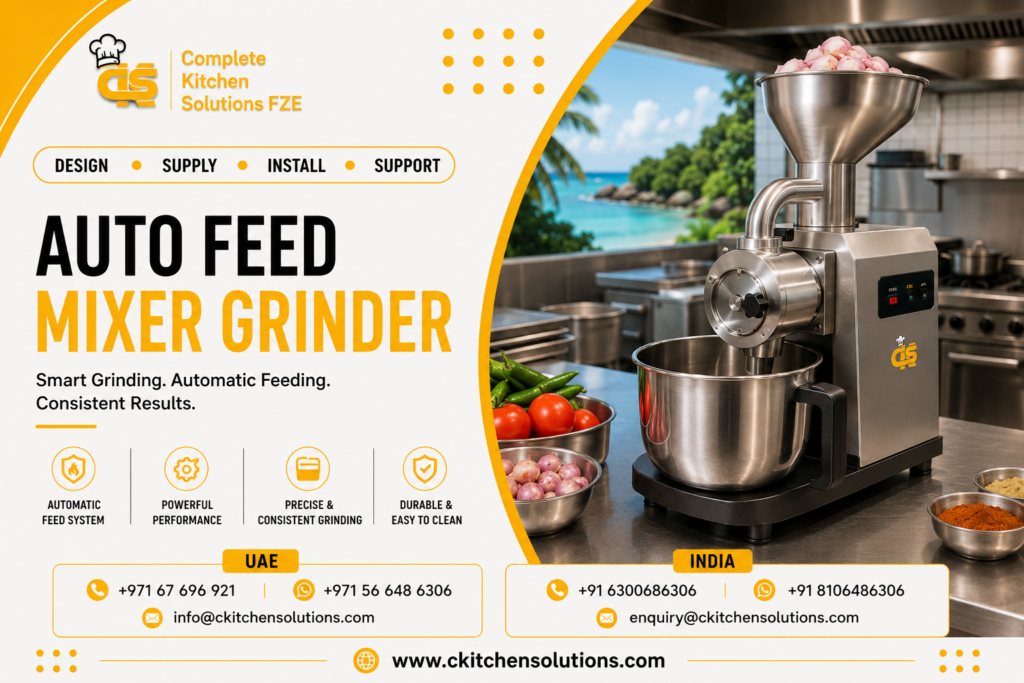 A promotional banner for "Complete Kitchen Solutions FZE" titled "Auto Feed Mixer Grinder: Smart Grinding. Automatic Feeding. Consistent Results," featuring a commercial stainless steel auto-feed grinding machine with a top funnel hopper loaded with shallots/onions, an attached collection mixing bowl, and bowls of fresh vegetables and ground spices on a prep counter against a tropical beach view.